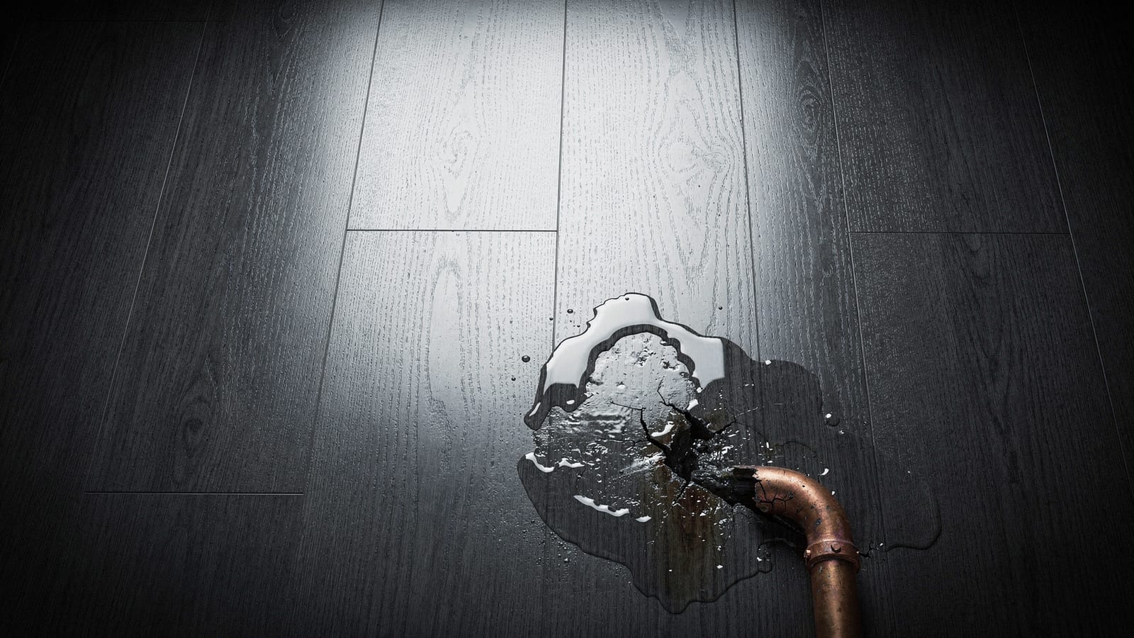 A cracked copper pipe leaking onto a dark hardwood floor